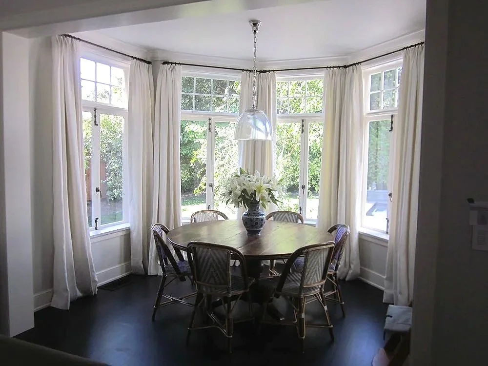 A round wooden dining table with six chairs around it, decorated with a large vase of white flowers. The table is in a bay window area with white curtains and multiple windows letting in natural light. A pendant light hangs above the table.