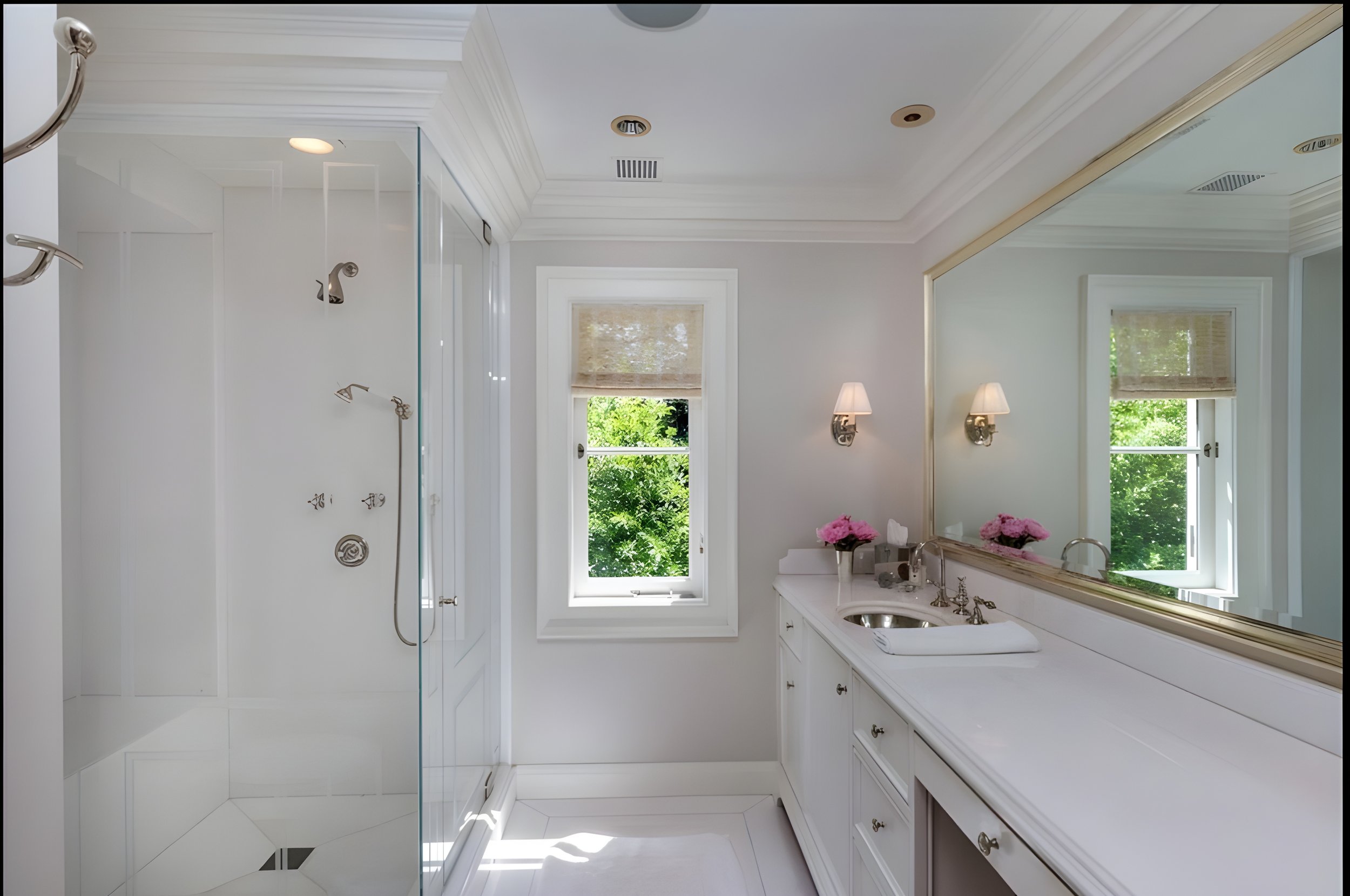 Bright bathroom with white cabinetry, a large mirror, two wall sconces, pink flowers, and a window overlooking green trees; a glass-enclosed shower is on the left side.