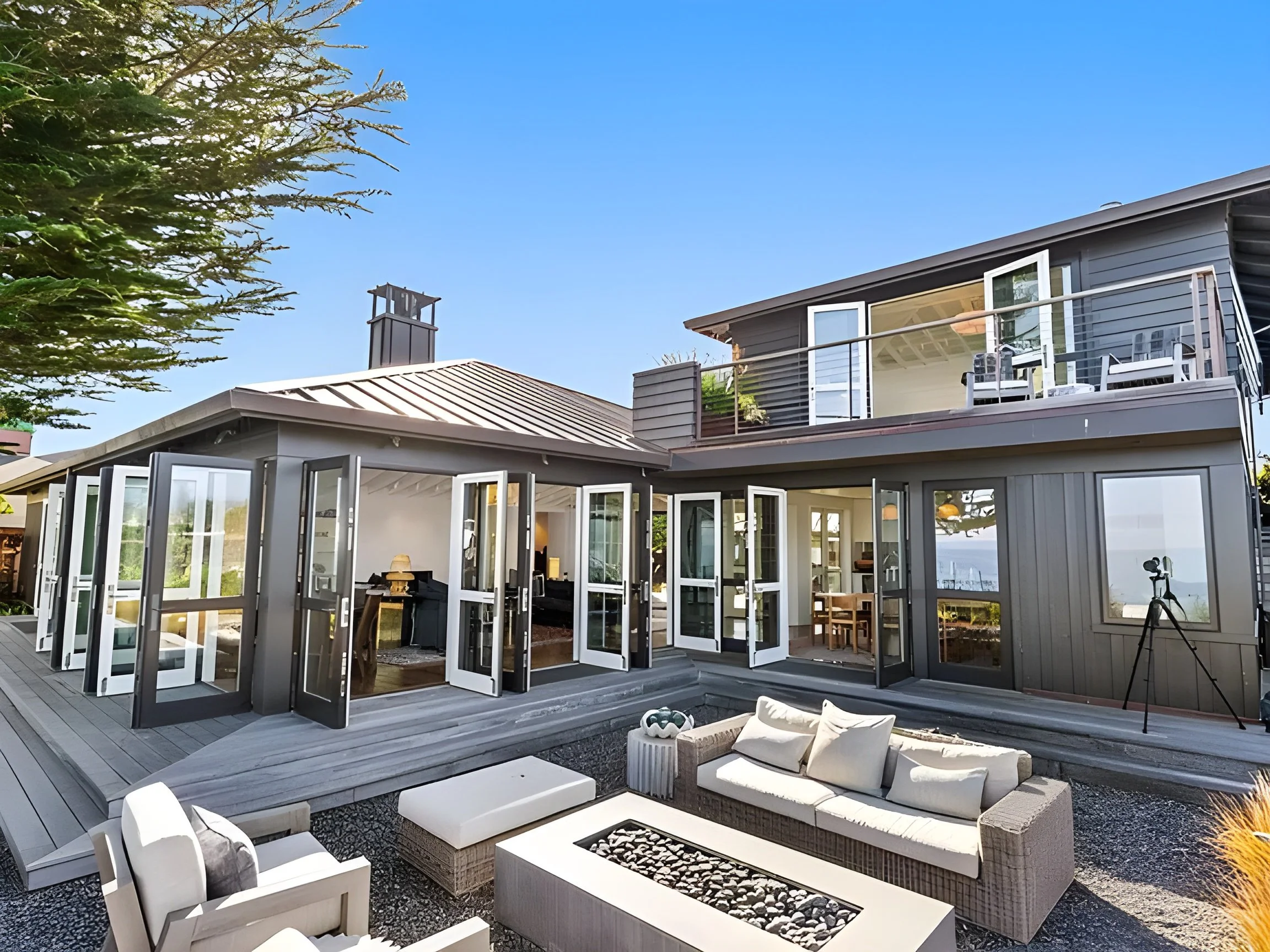 Modern house with multiple glass-paned doors and windows, outdoor seating area, and a balcony, during daytime with clear sky.
