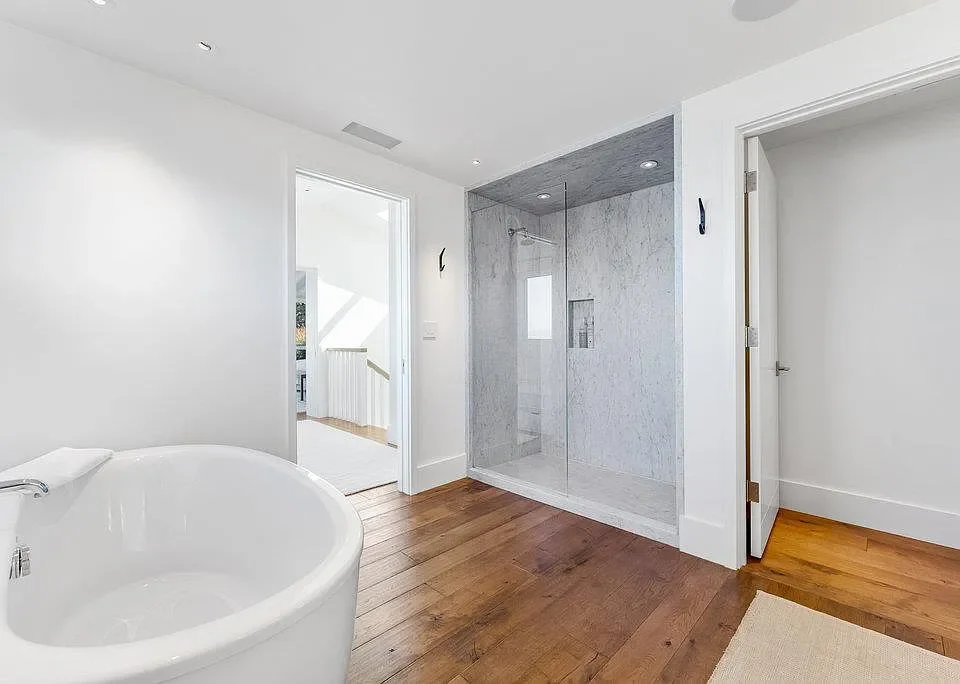 Bathroom with a white bathtub, walk-in shower with glass door and marble walls, wooden flooring, and a doorway leading to a staircase.