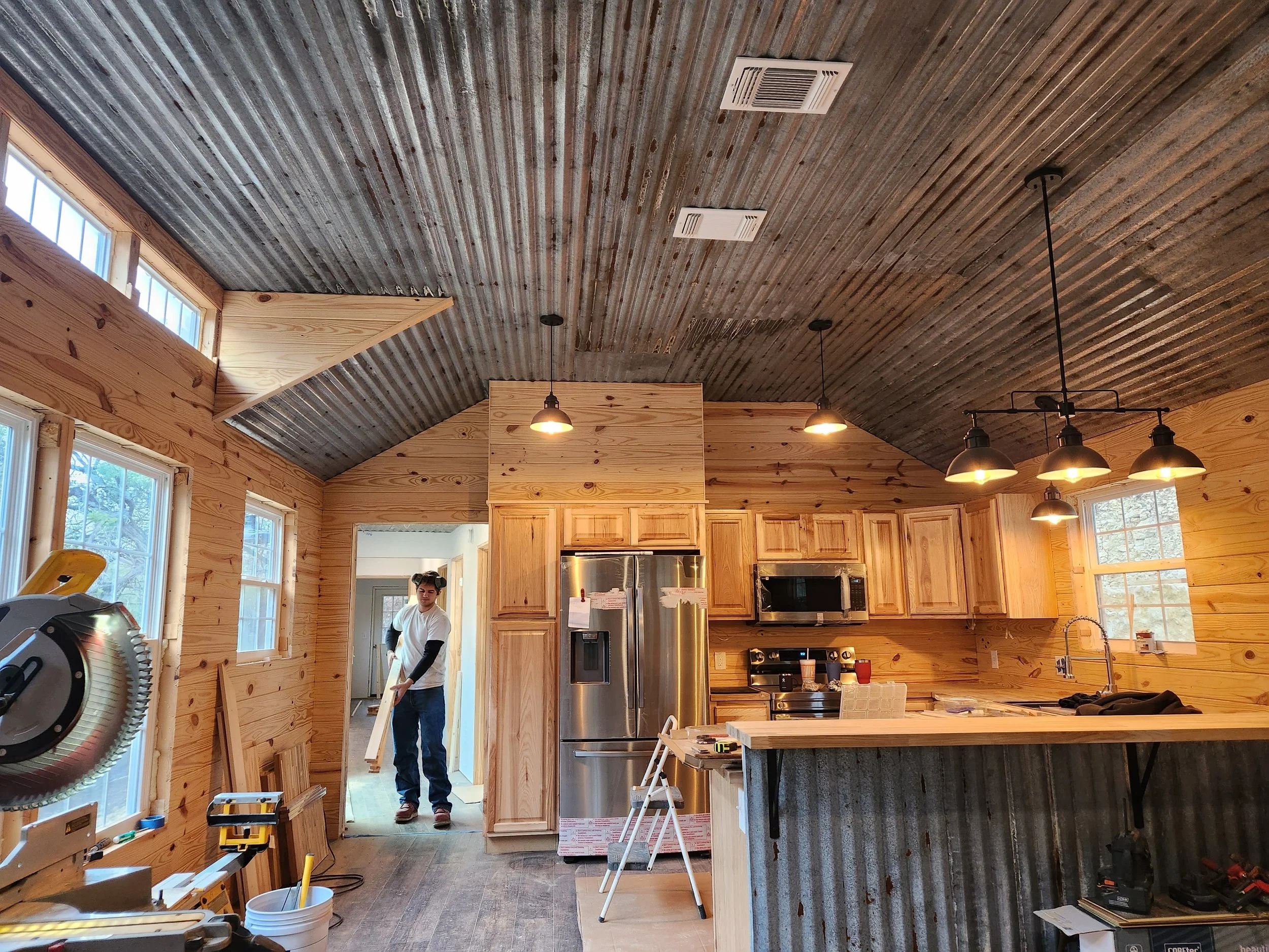 A person holding a piece of wood in a partially finished kitchen with wooden walls, a roof with exposed corrugated metal, and hanging pendant lights.