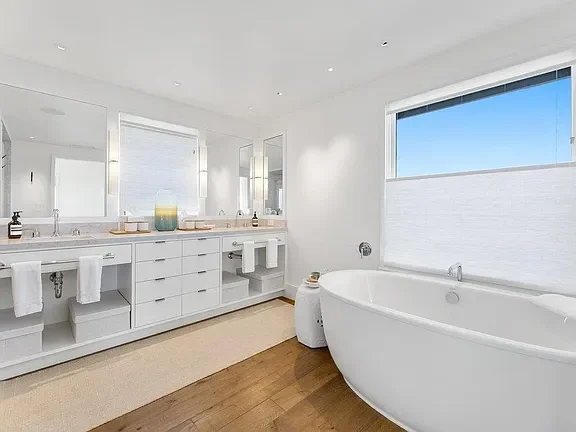 Modern bathroom with white double vanity, large mirror, and a barrel-shaped bathtub by a window.