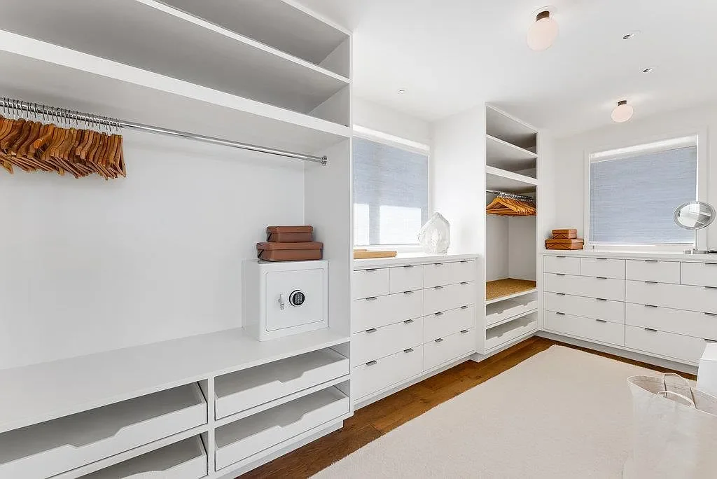 Empty walk-in closet with white built-in shelves, drawers, and hanging space, with brown hangers and folded brown boxes, hardwood floor, and two windows with blinds.