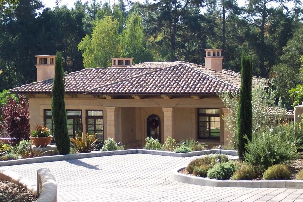 Front view of a house with a tiled roof, surrounded by trees and landscaping, including tall cypress trees and drought-tolerant plants.