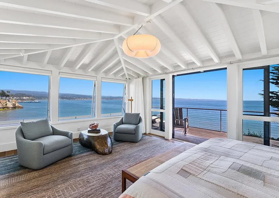 A bright bedroom with large windows and a sliding glass door overlooking the ocean. The room has a bed, two armchairs, a coffee table, and a view of a deck with outdoor seating.