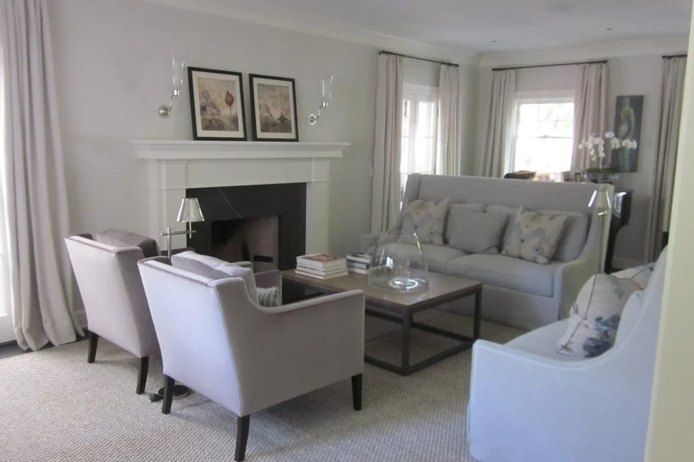 Living room with a fireplace, framed artwork, cream-colored armchairs and sofa, coffee table with books and a glass dome, beige curtains, and decorative pillows.