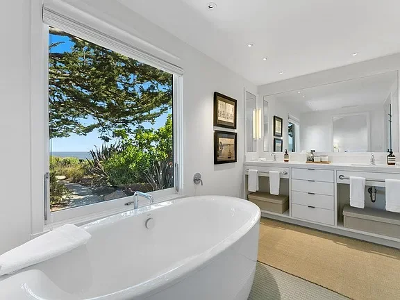 Modern bathroom with white bathtub, large window showing outdoor trees and sky, double sink vanity, mirror, and framed artwork.