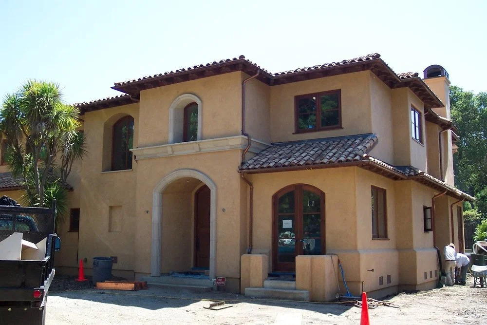 A two-story house under construction with a stucco exterior, red tile roof, and multiple arched windows and doors, situated in a tropical setting with palm trees.