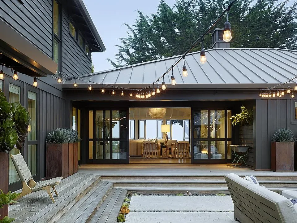 Exterior view of a modern house deck with string lights, potted plants, outdoor seating, and an indoor dining area visible through large glass doors.
