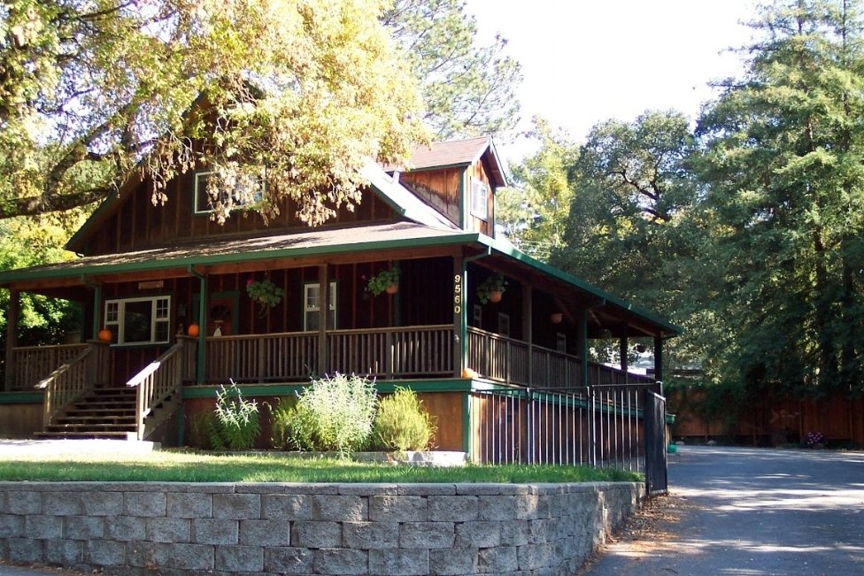 A two-story wooden house with a porch decorated with pumpkins, surrounded by trees, with a driveway on the right side.