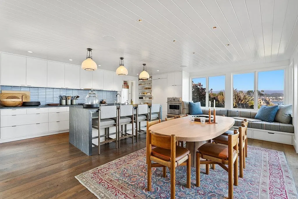 Open kitchen and dining area with white cabinets, a blue tiled backsplash, a kitchen island with bar stools, a wooden dining table with chairs, and large windows offering a view outside.