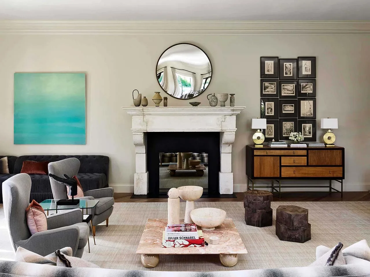 A living room with a white fireplace, a round mirror above, and various decorative items on the mantel. To the right, there is a wooden sideboard with lamps and framed photos. On the left, there is a black sofa, a gray armchair with pink pillows, and