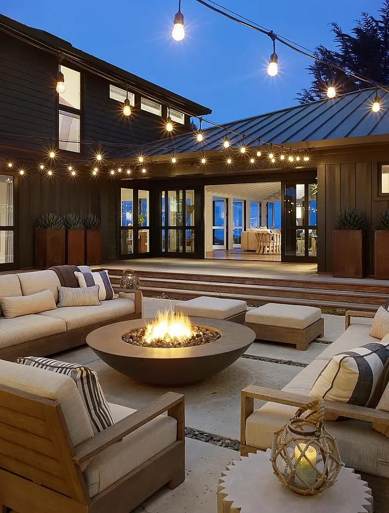 Outdoor patio with beige seating around a fire pit, string lights overhead, and a modern house with large windows and a patio in the background at dusk.