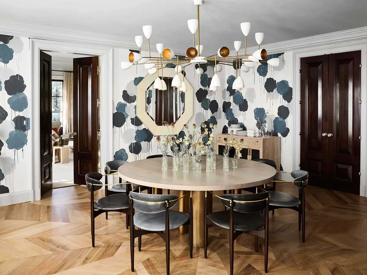A dining room with a round table, black chairs, a modern chandelier, a mirror on the wall, and a wall decorated with blue, black, and gray abstract patterns.