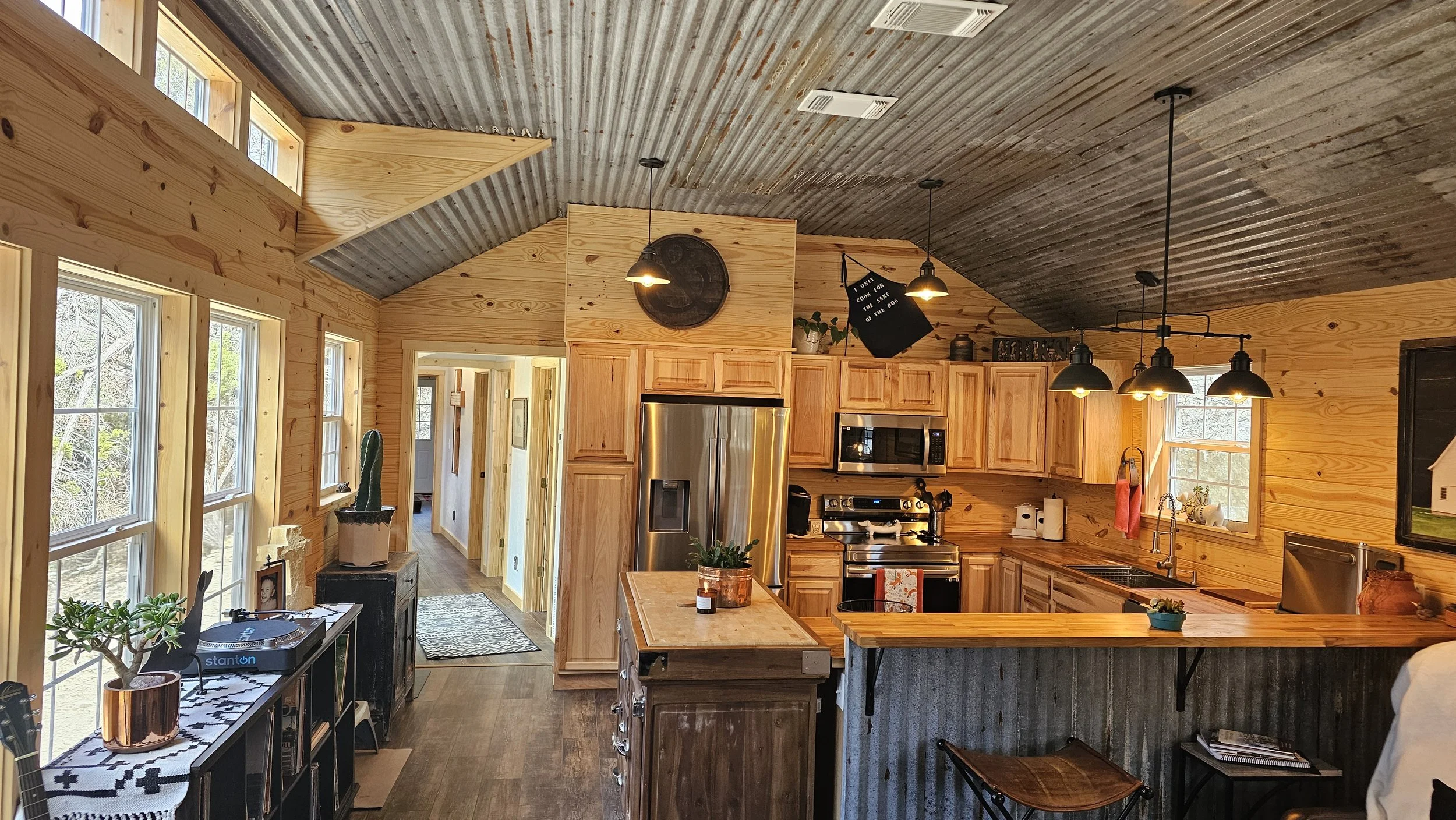 A rustic kitchen with wood-paneled walls and a corrugated metal ceiling, featuring stainless steel appliances, a small kitchen island, and pendant lighting, with three large windows letting in natural light.