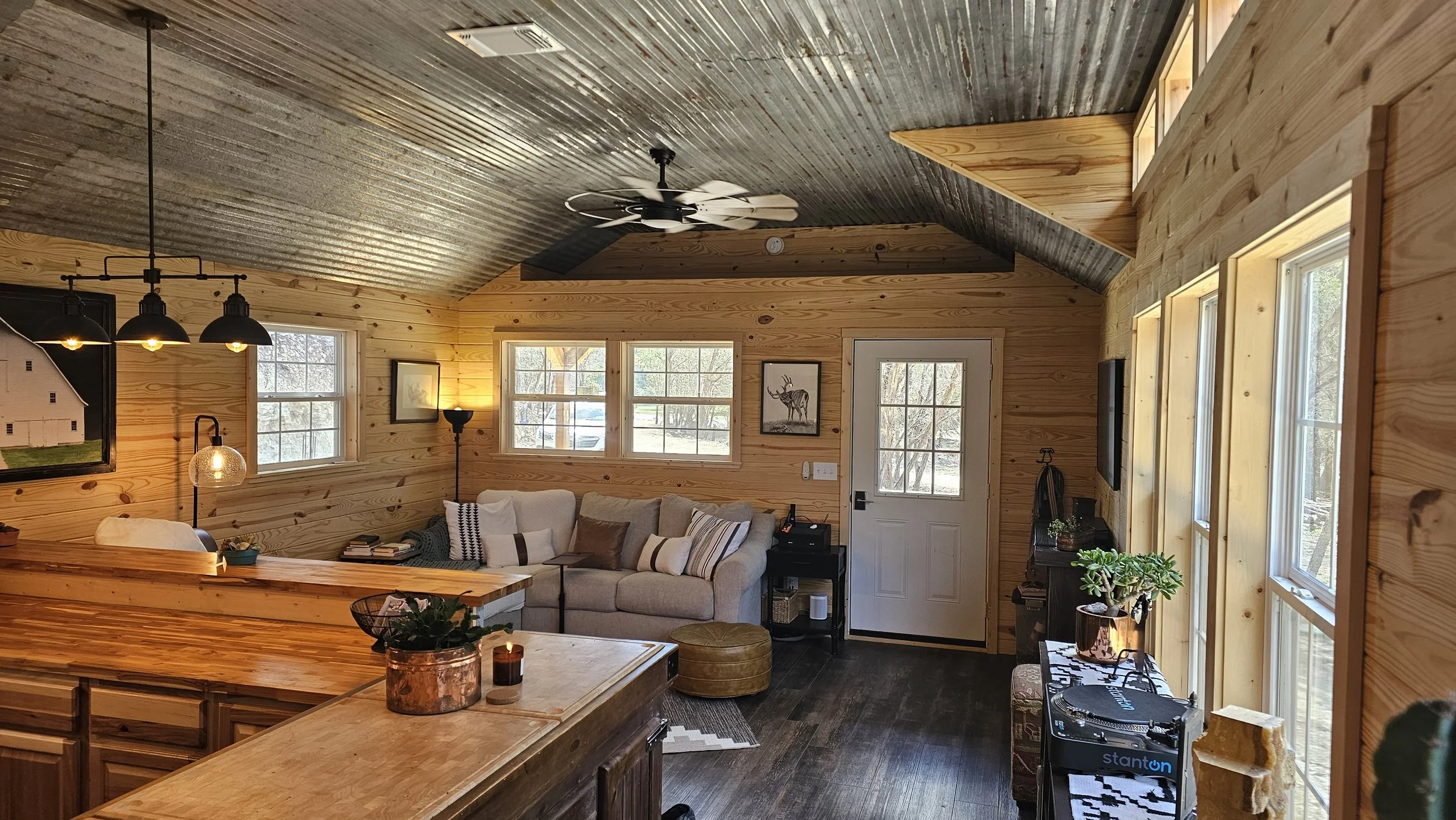 Cozy living room with wood-paneled walls, a gray sofa with pillows, a black side table, large windows, and a ceiling fan.