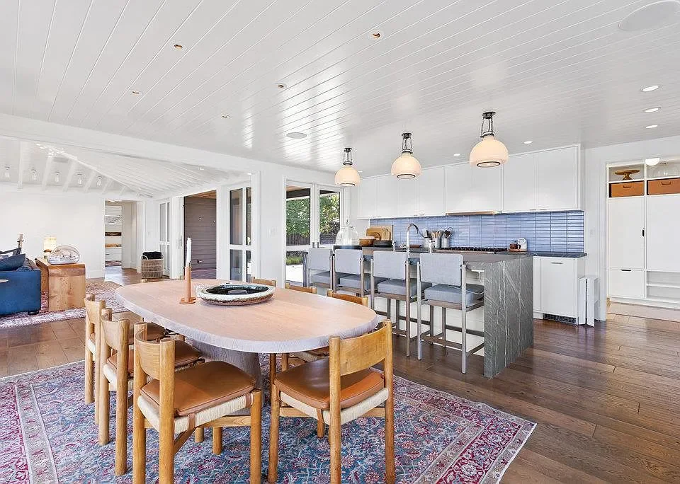 Open-concept kitchen and dining area with white cabinetry, light blue backsplash, island with bar seating, wooden dining table with chairs, and hardwood floors