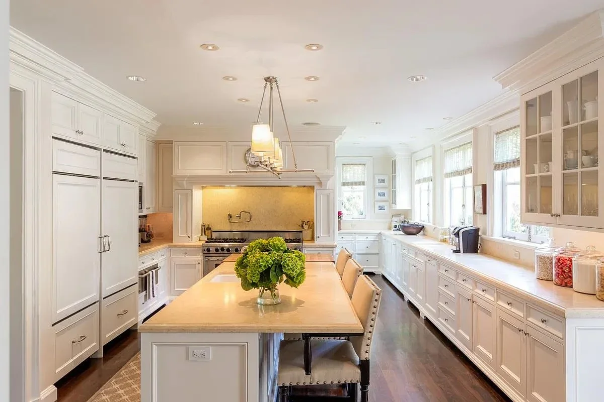 Bright kitchen with white cabinets, large windows, a central island with a flower arrangement, and various appliances and jars on the counters.