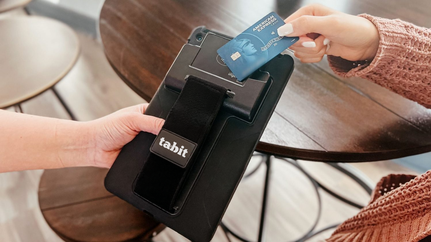 Person swiping a blue American Express credit card at a contactless payment terminal, with a black tabit bag hanging on it, on a wooden table.