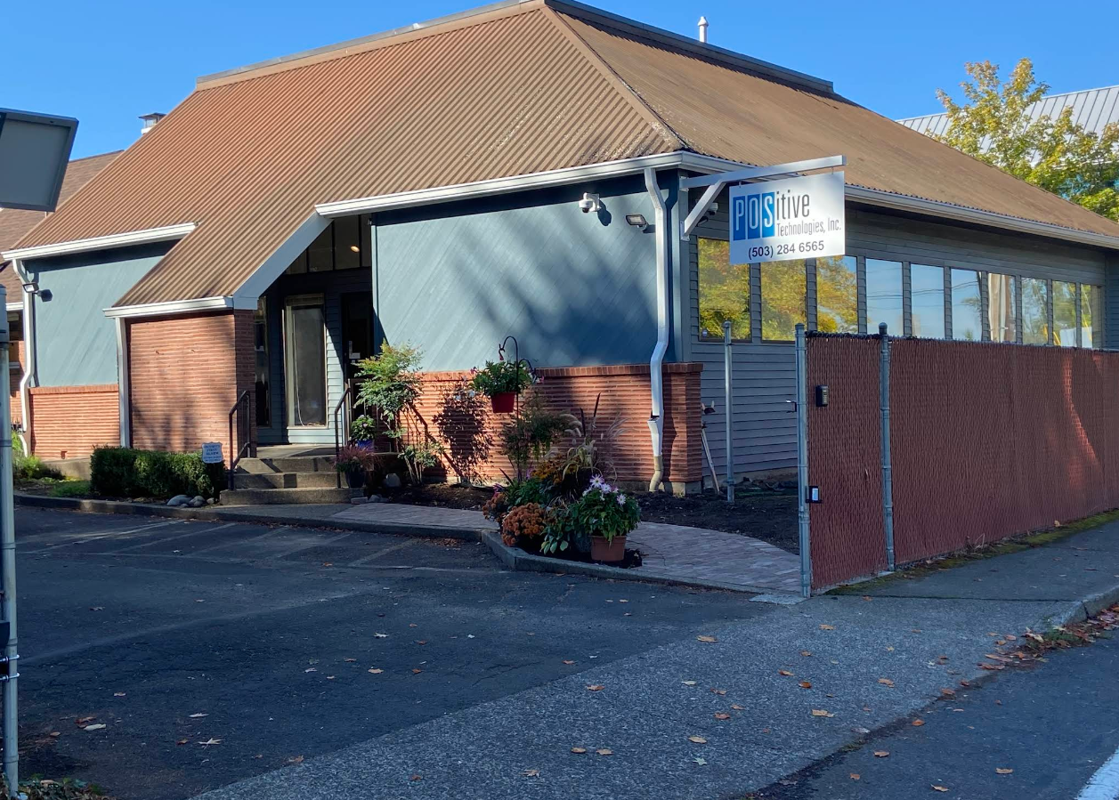 A building with a brown sloped metal roof, blue exterior walls, and large windows on the right side. There are plants and flowers in front of the entrance, which has a small staircase and a glass door. A sign for Posiitive Technologies, Inc. is attached to the building near the corner.