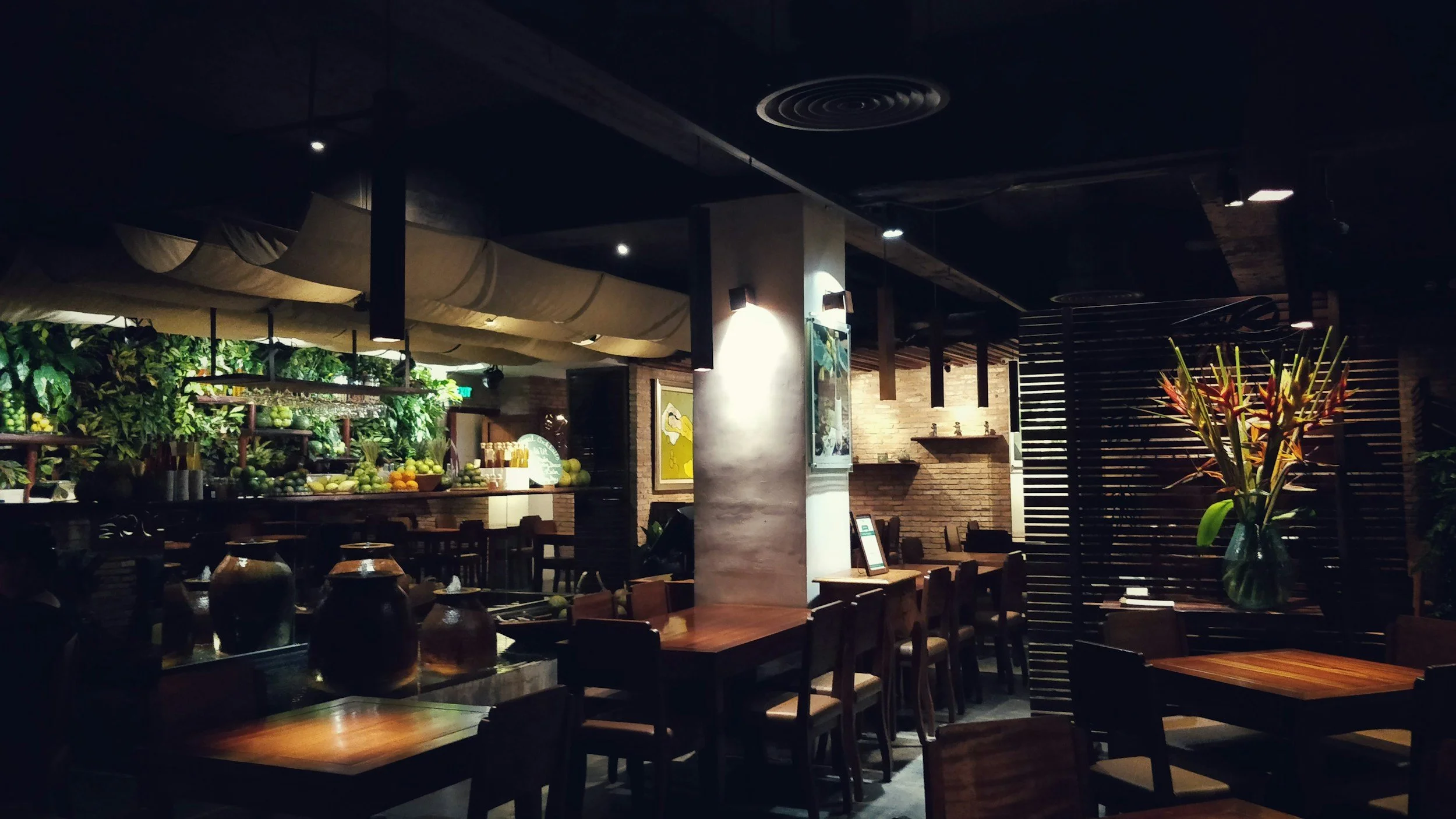 Interior of a restaurant with wooden tables and chairs, decorative vases, lush green plants, and brick walls, dimly lit for a cozy atmosphere.