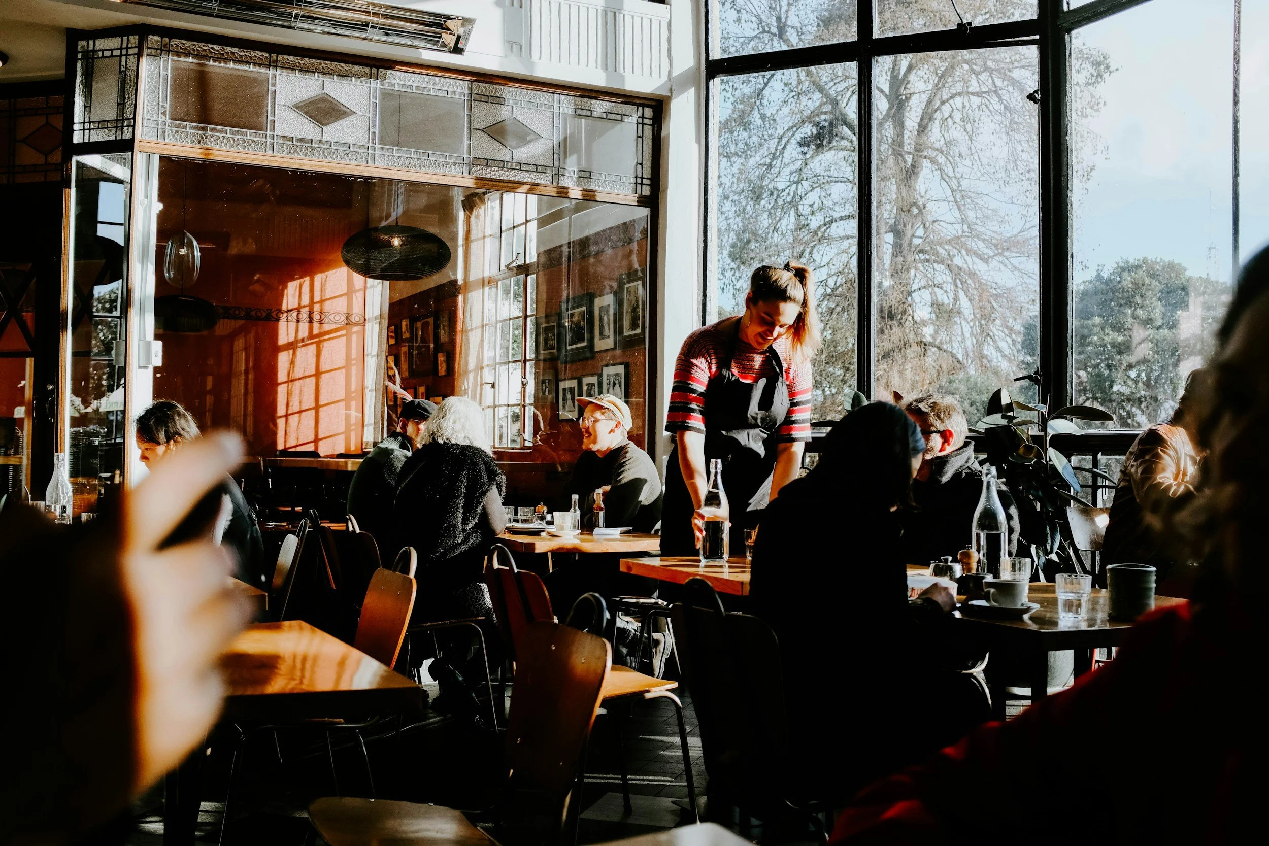 A busy restaurant interior with large windows letting in sunlight, showing people dining and a waitress serving at a table.