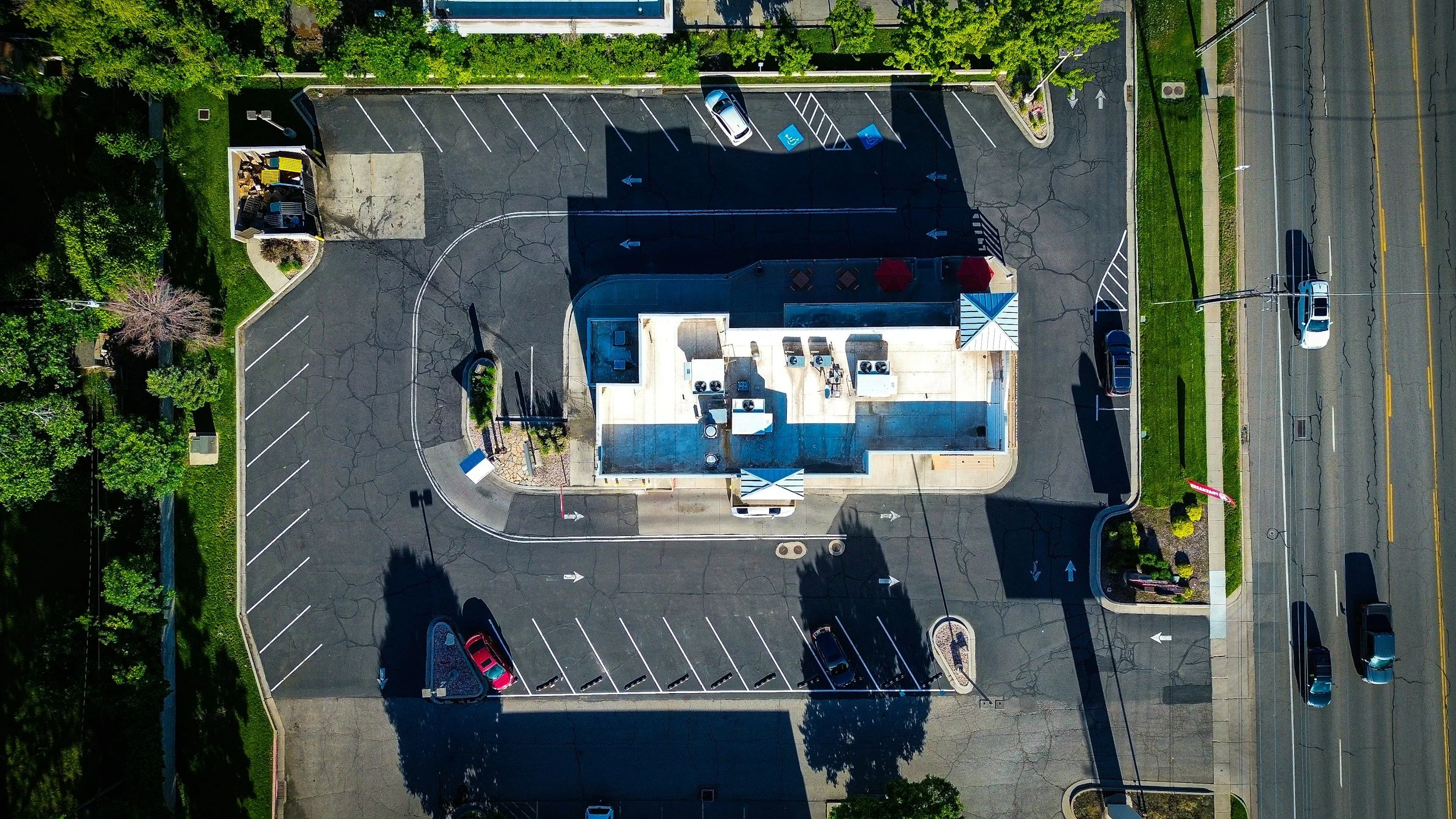 An aerial view of a commercial building with a parking lot, surrounded by trees and a street with cars passing by