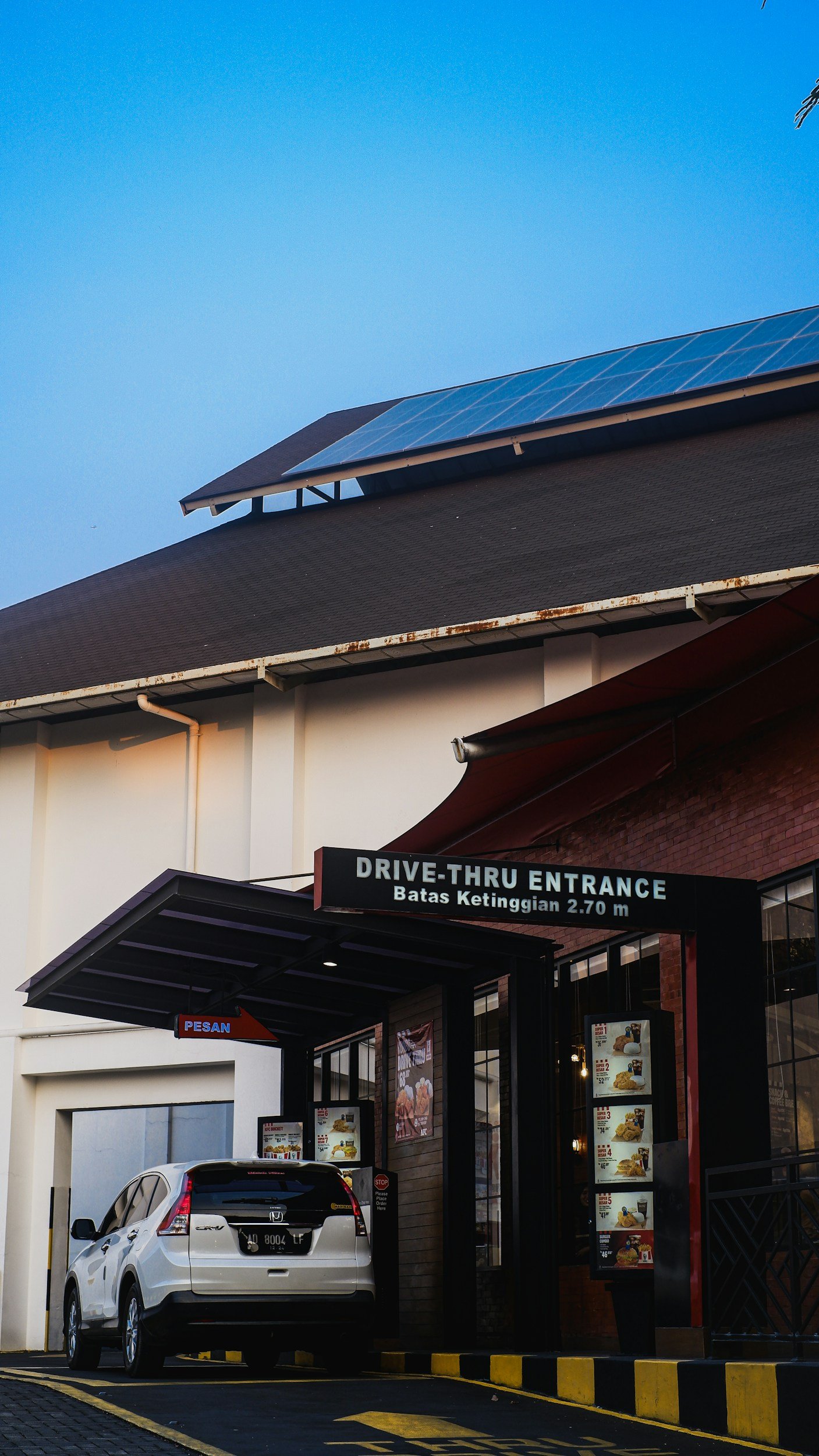 View of a drive-thru restaurant entrance with a white car parked, a sign indicating the drive-thru entrance, and menu boards visible. The building has a dark roof with solar panels and a clear blue sky.