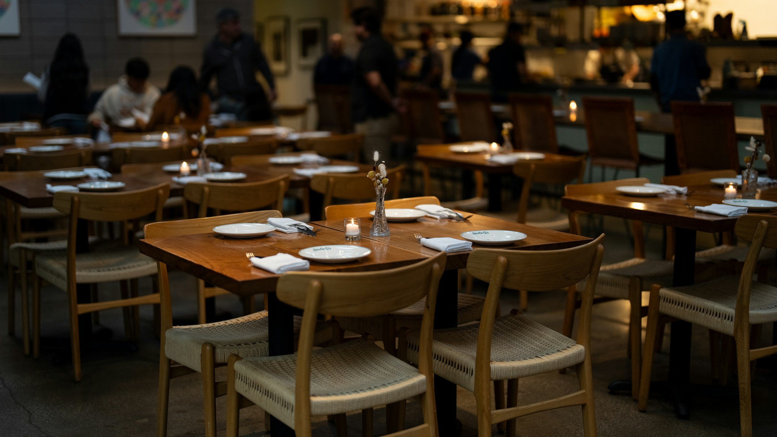Empty restaurant tables set with white plates, napkins, utensils, small candles, and vases with flowers, with a few customers and staff in the background.