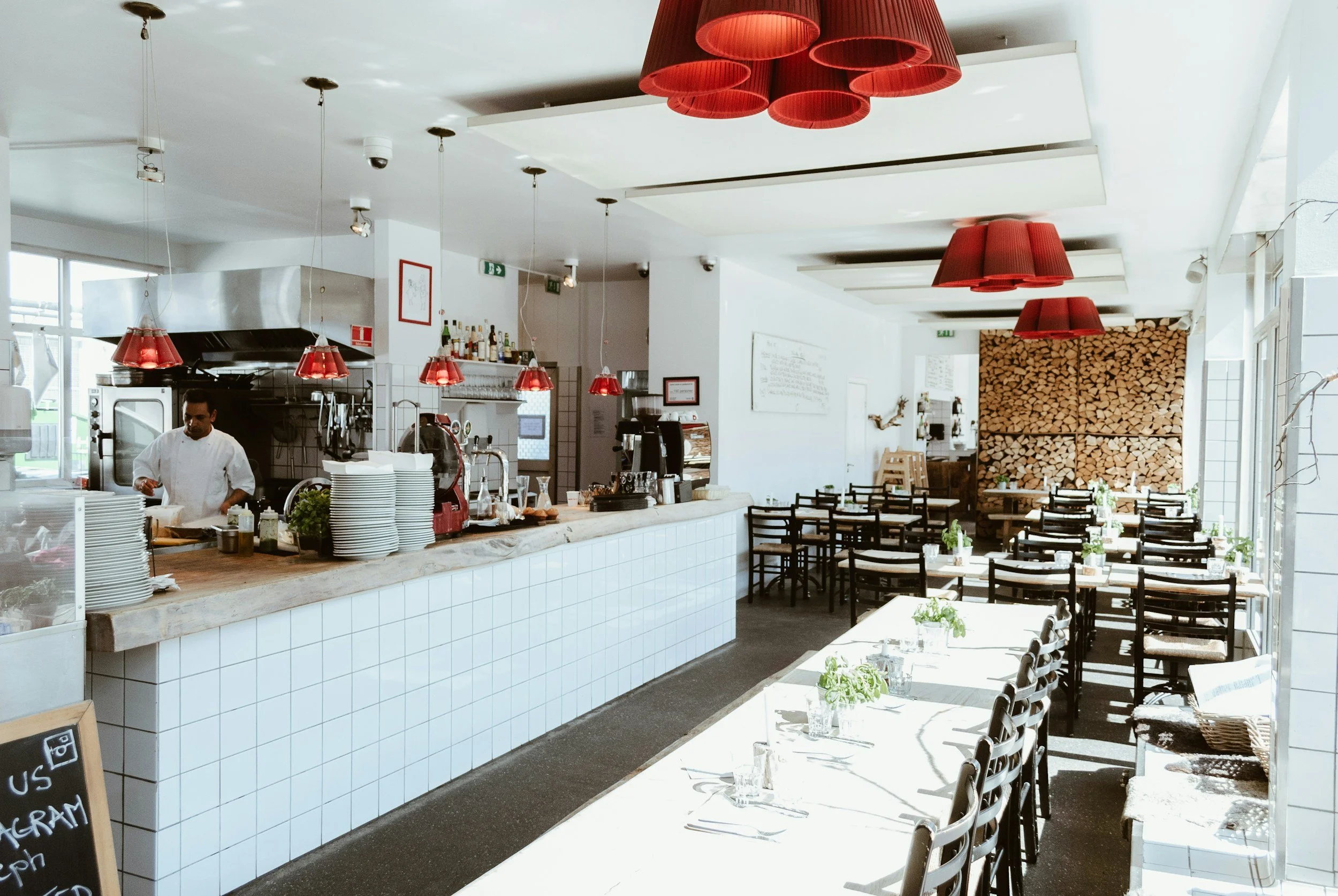 Interior of a bright, modern restaurant with a long white-tiled bar, red pendant lamps, a kitchen station with a chef, and neatly set tables with plants and glasses.