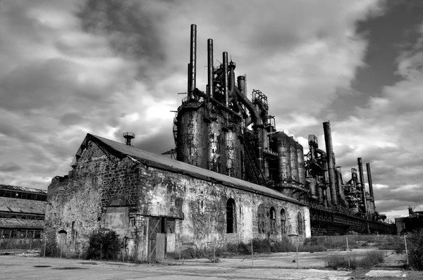 An abandoned, rusted industrial factory with multiple tall smokestacks and old brick buildings, under a cloudy sky.
