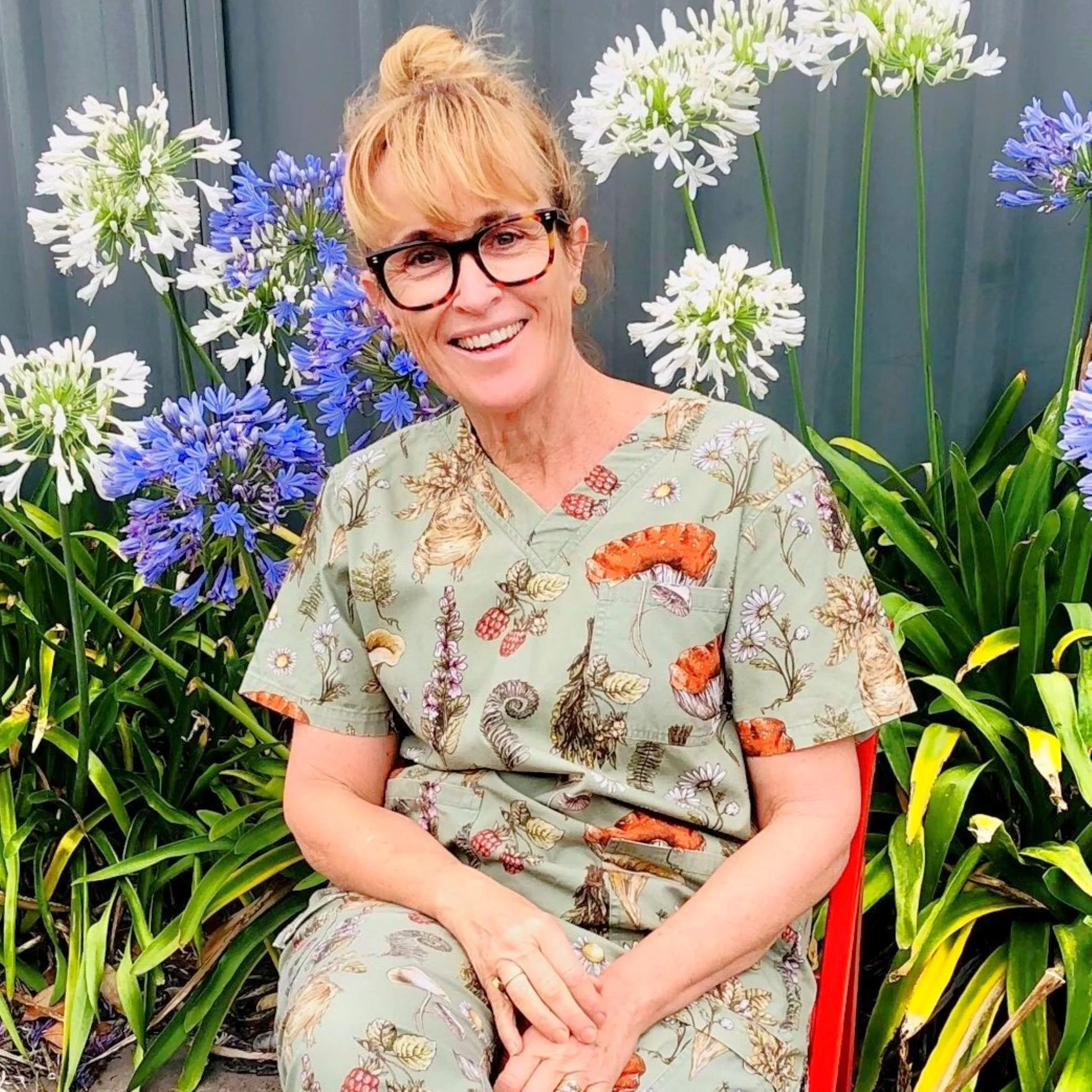 A smiling woman with glasses and blonde hair tied in a bun, wearing scrubs with a nature print, sitting in front of blooming white and purple flowers.