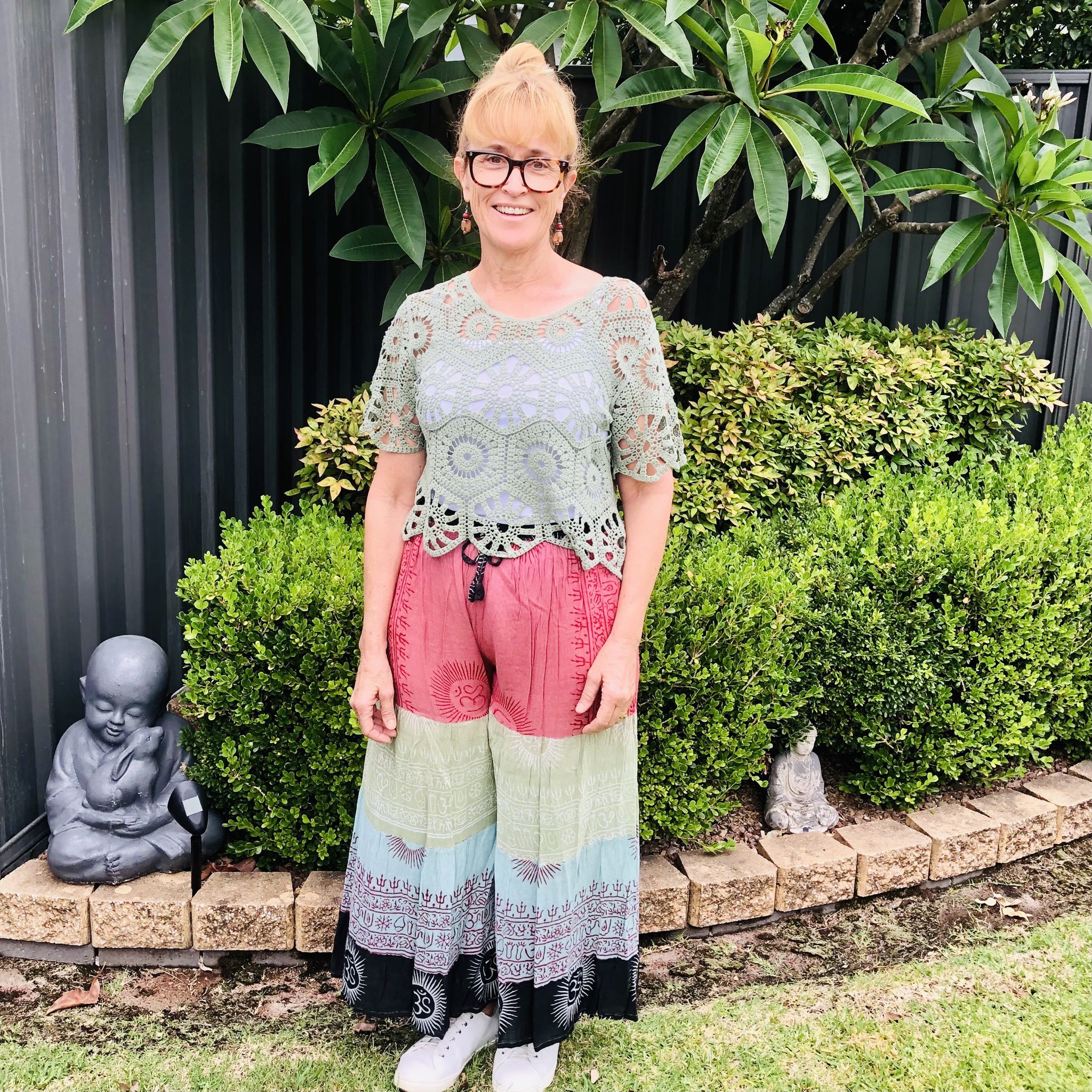 A woman with blonde hair in a bun, wearing glasses, a light green crochet top, and colorful wide-leg pants, standing outdoors in front of green bushes and statues of a seated Buddha and a meditating figure.