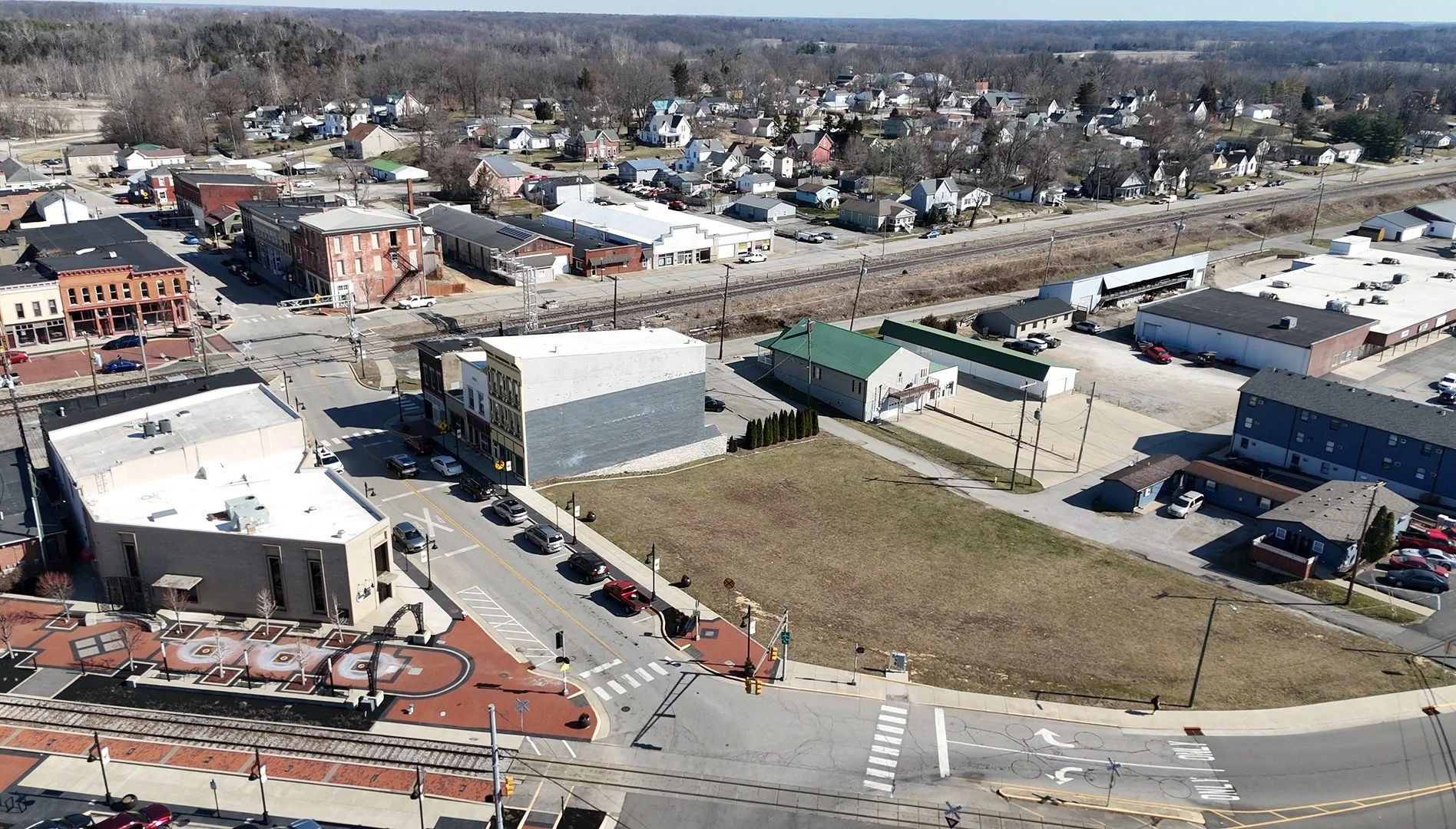 Aerial view of a small town with a railway line, buildings, streets, and sidewalks. There are parked cars and some moving vehicles, with a mix of residential and commercial areas. The landscape extends to a lightly wooded area in the background.