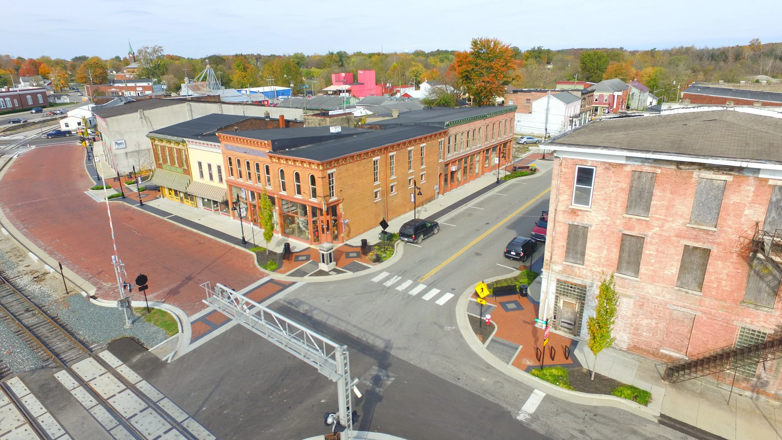Aerial view of a small town with brick buildings, a street with parked cars, railroad tracks, and autumn trees in the background.