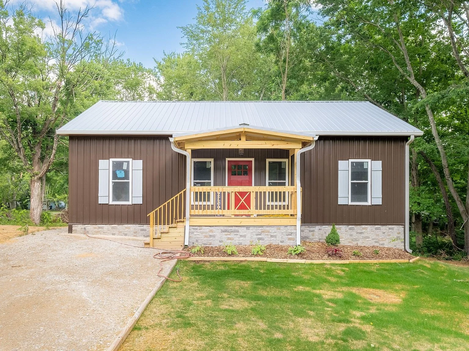 Newly constructed small house with dark brown vertical siding, white window shutters, stone foundation, and a metal roof. It features a small front porch with a wooden railing and a red front door. The house is surrounded by a green lawn and trees.