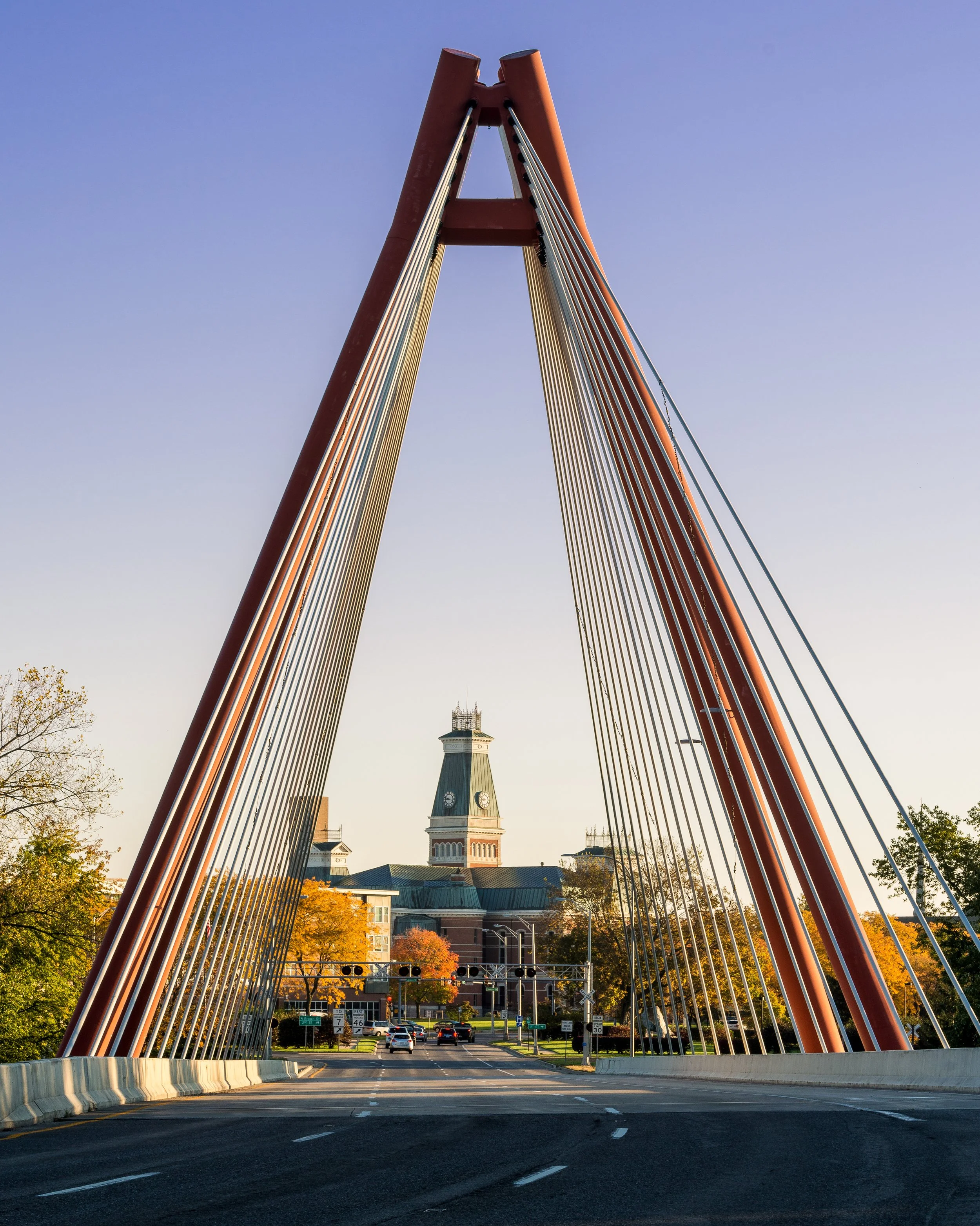 View of a bridge with orange supports and cables, leading to a historic courthouse with a clock tower in the background during fall, with autumn trees and cars on the road.