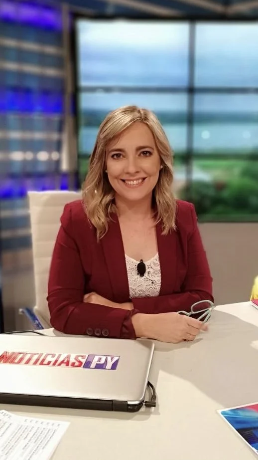 Mujer sonriendo en un estudio de noticias, lleva blazer rojo y está frente a una laptop que dice "Noticias PV".