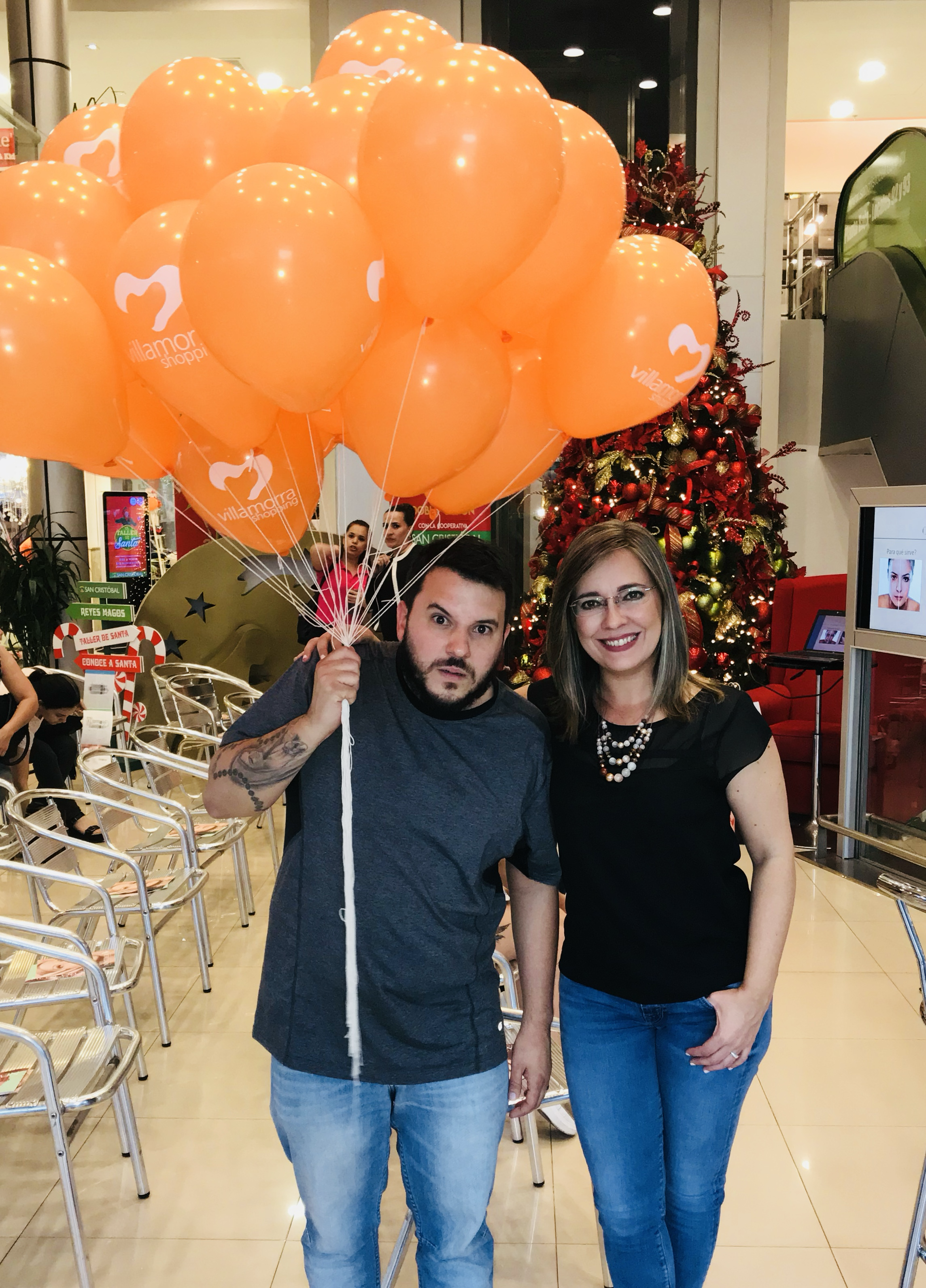 Una pareja posando con globos naranjas con logotipo de Villamora Shoppi en un centro comercial decorado con árbol de Navidad y muebles de cafetería.