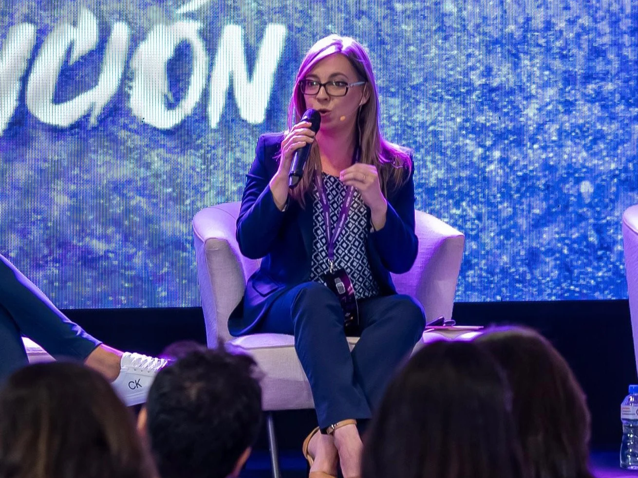 Una mujer con gafas y cabello rubio, vestida con traje azul oscuro, está sentada en una silla y hablando en un micrófono en un escenario con fondo azul con letras y efectos visuales.
