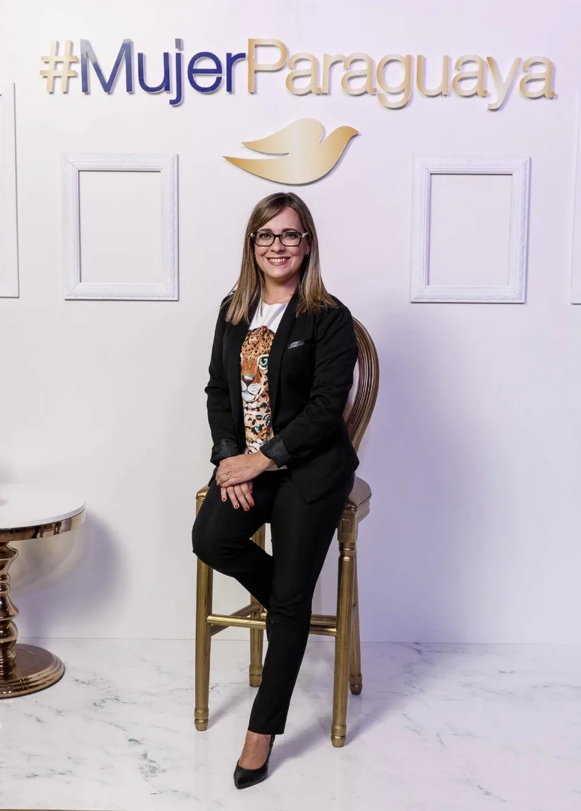 Mujer sonriendo, vestida con traje negro y blusa con diseño de jaguar, sentada en una silla dorada frente a fondo blanco con el texto #MujerParaguaya y un símbolo de paloma dorada.