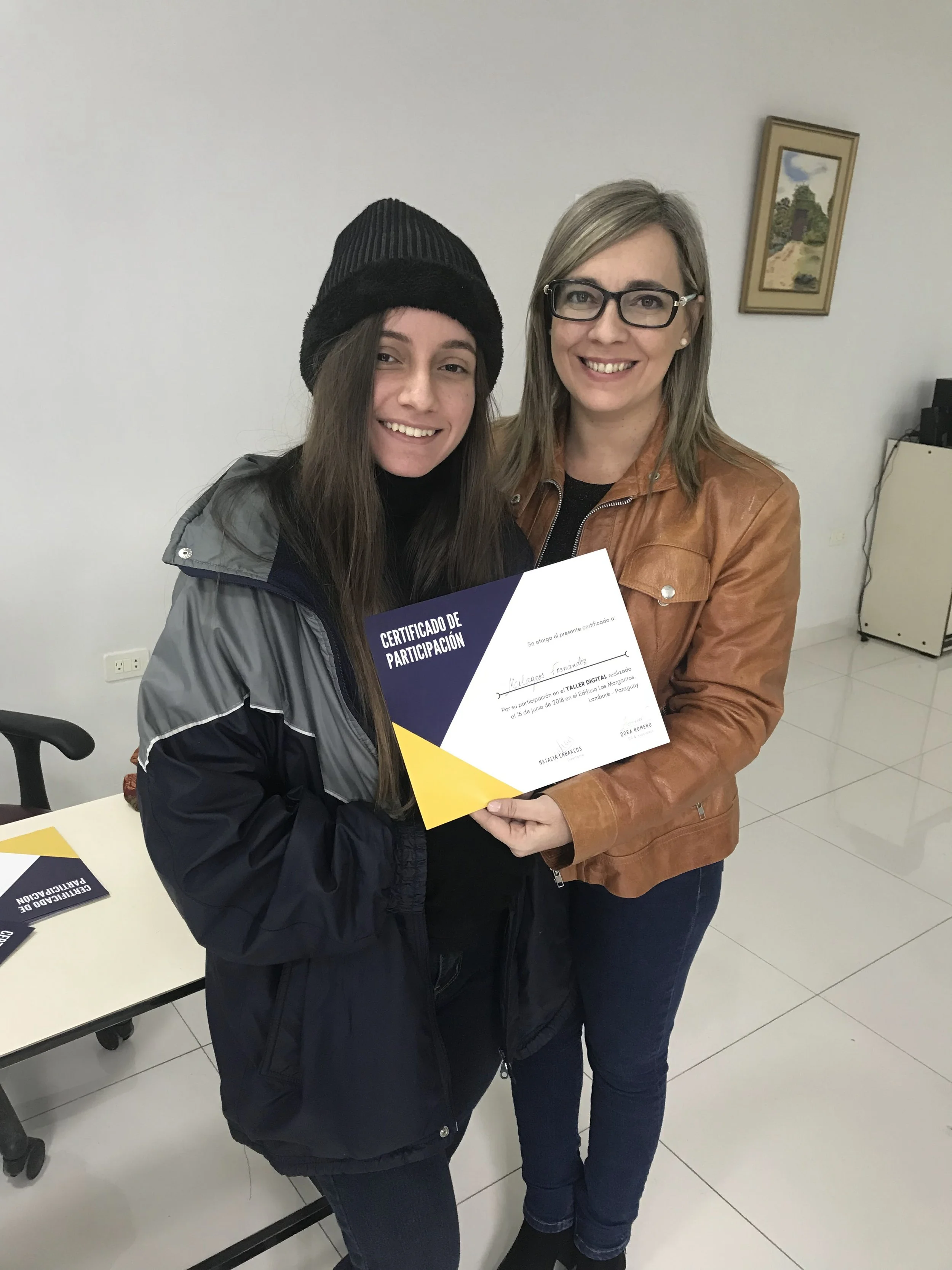 Mujer joven recibiendo un certificado de participación de parte de otra mujer en un ambiente interior con paredes blancas y cuadro en la pared.