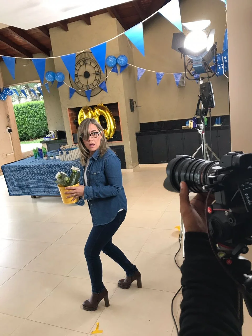 Mujer con gafas, en una celebración, sosteniendo una maceta con flores, en un espacio decorado con banderines azules, globos y un reloj grande en la pared, con equipo de filmación frente a ella.
