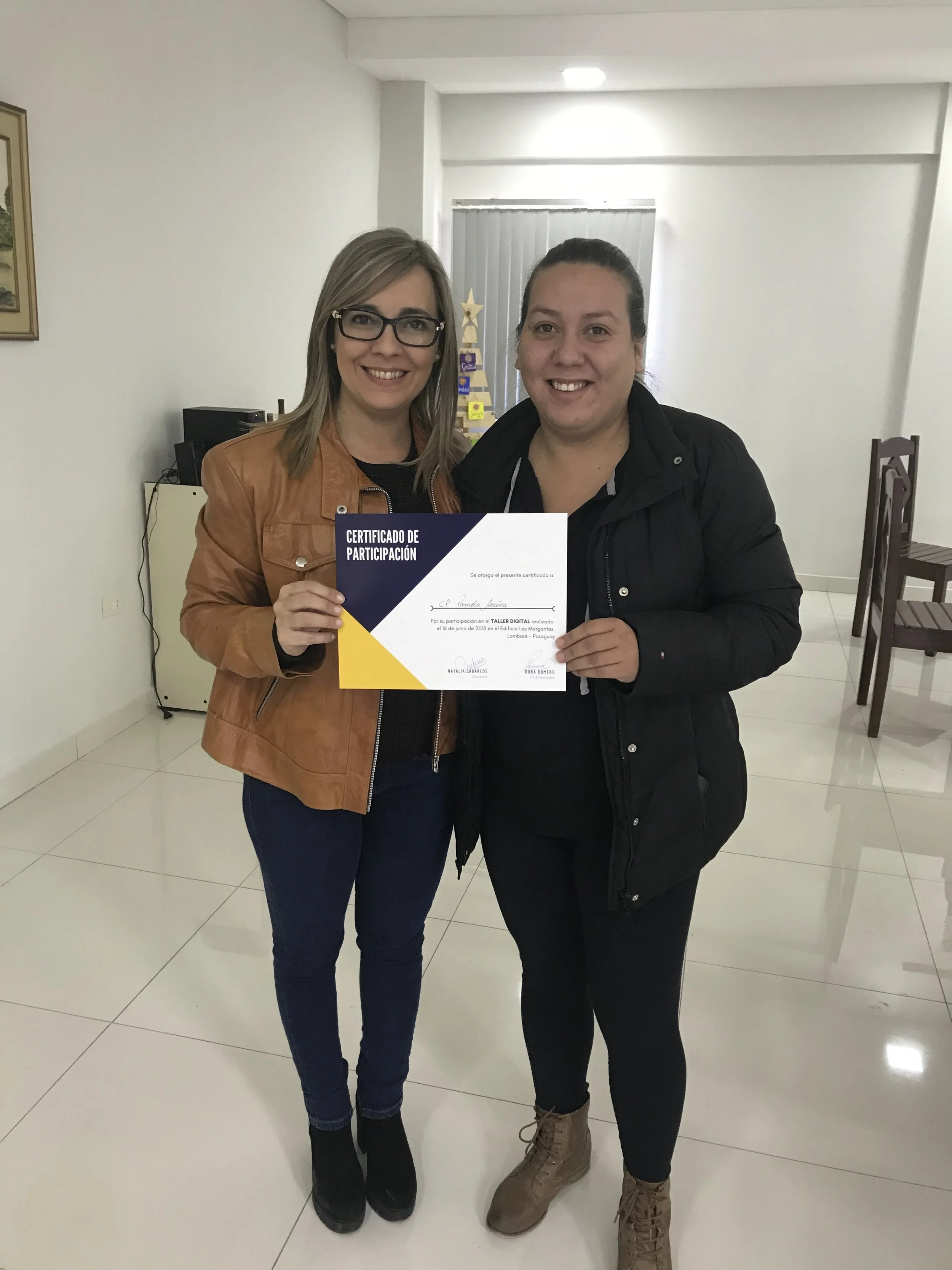 Dos mujeres sosteniendo un certificado de participación, una con gafas y chaqueta de color marrón claro, la otra con cabello recogido y chaqueta negra en un salón con sillas y decoraciones.