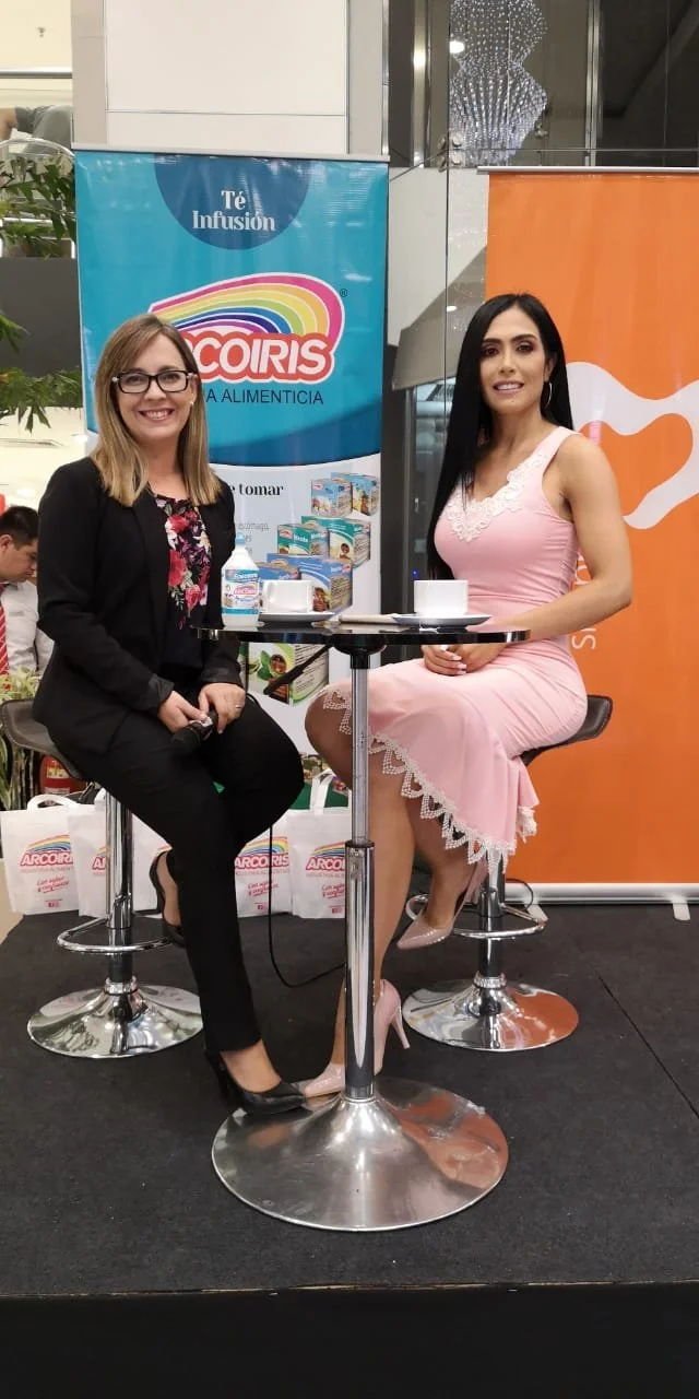 Dos mujeres sentadas en taburetes junto a una mesa, una con gafas y ropa negra con flores y la otra con vestido rosa, en un evento promocional con banners de Arcoiris y Té Infusión.
