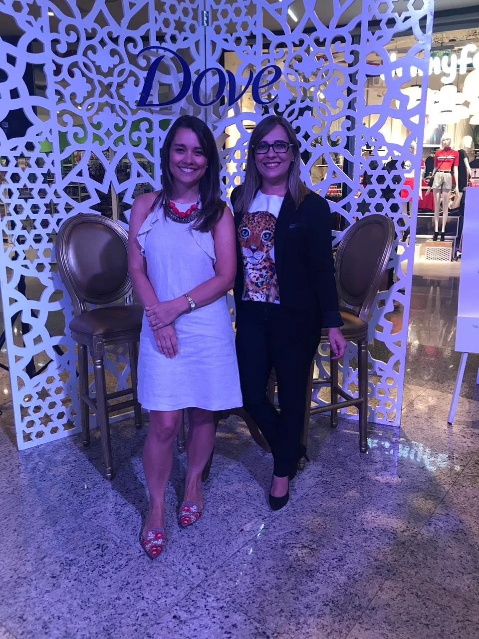 Dos mujeres posando delante de una estructura decorativa blanca con la palabra 'Love' escrita, en un lugar que parece una tienda o centro comercial, con maniquíes y ropa visible al fondo.