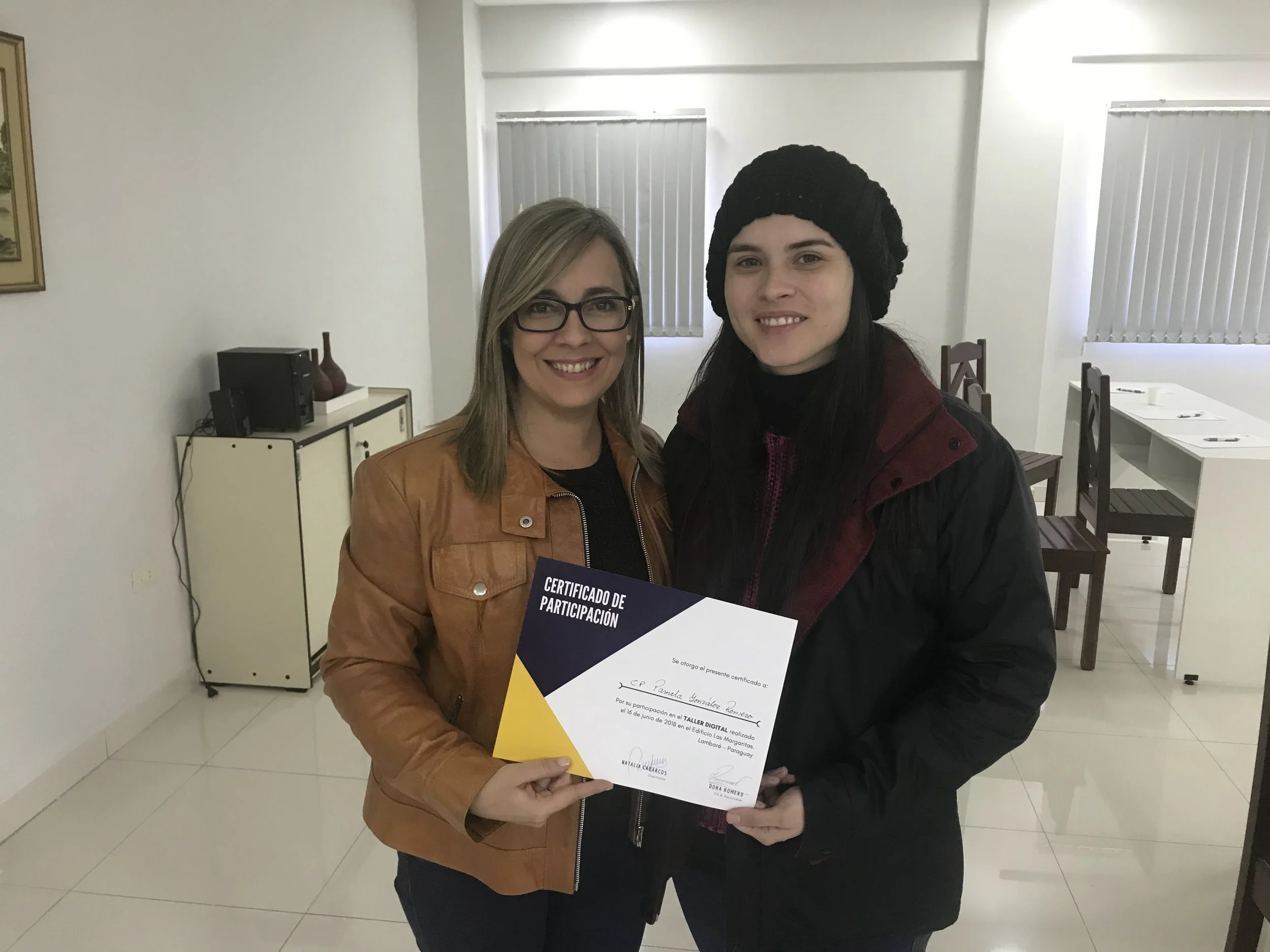 Dos mujeres sonriendo mientras sostienen un certificado de participación en una sala con sillas y ventanas con persianas verticales.