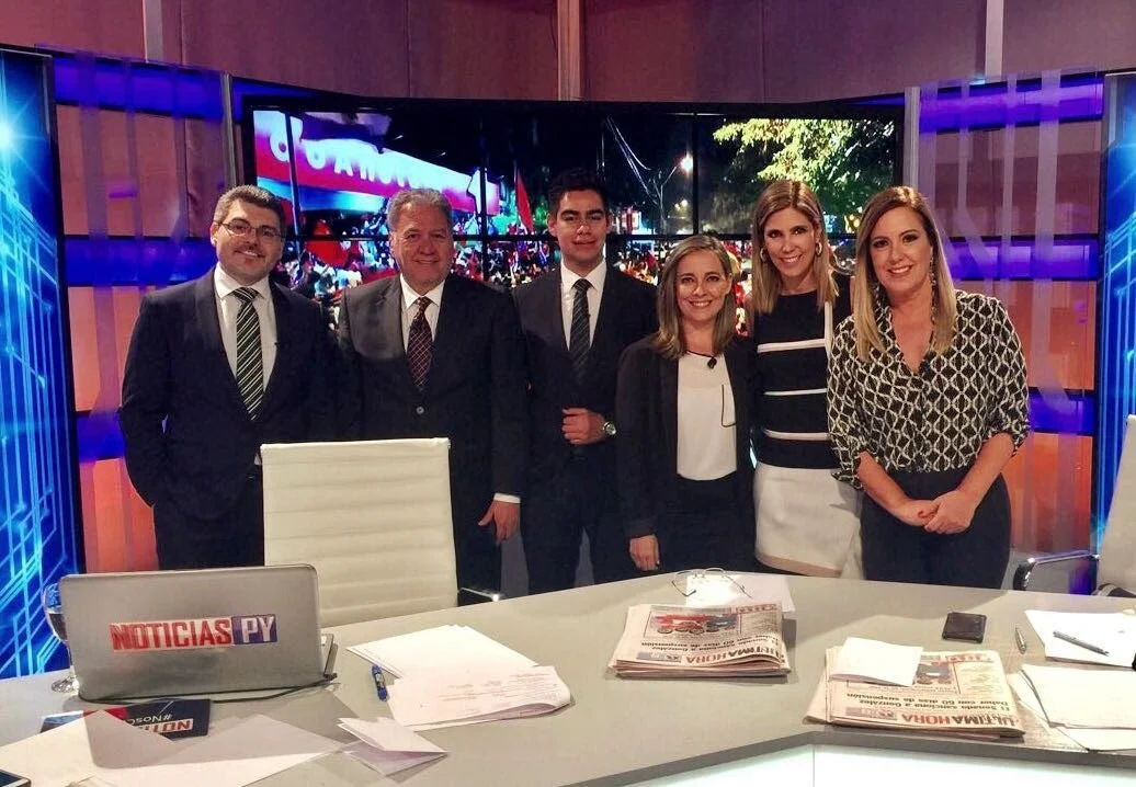 Grupo de seis personas en una sala de televisión, vestidos formalmente, frente a una mesa con periódicos y una computadora portátil que dice 'Noticias PY'.