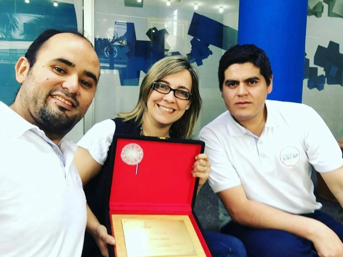 Tres personas sentadas, una mujer en el centro sosteniendo una placa de reconocimiento dentro de una caja roja, rodeada por dos hombres. Todos sonrientes, en un ambiente de oficina con decoraciones azules en el fondo.