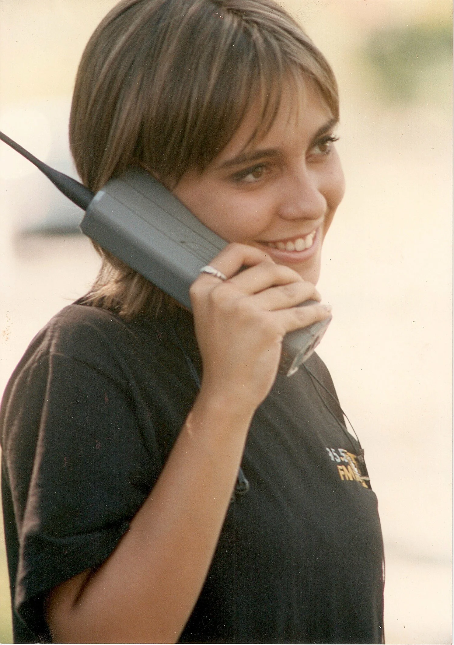 Mujer sonriendo y hablando por teléfono móvil, lleva una camiseta negra y mochila, cabello castaño corto.